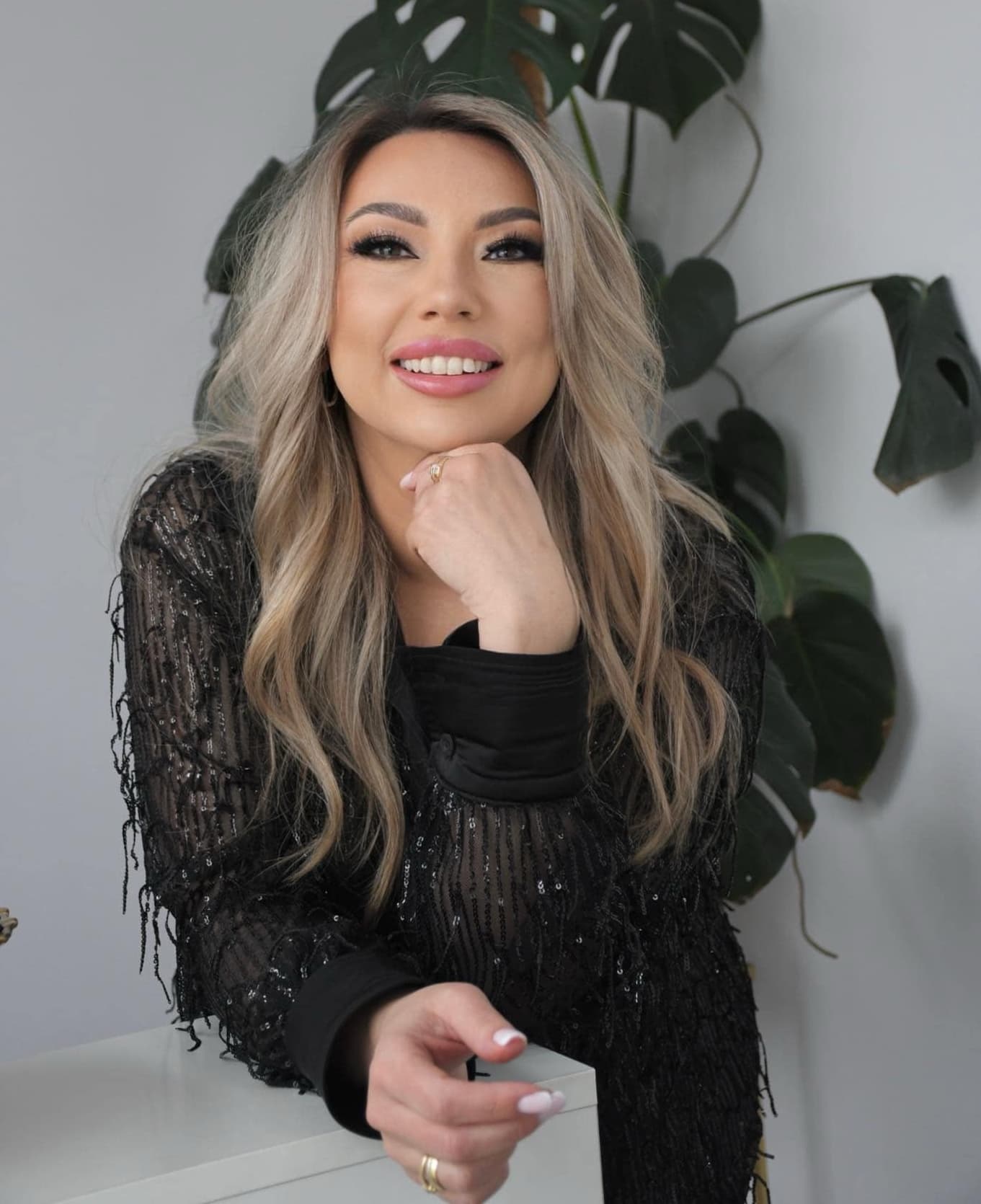 Olga Noszczyk uśmiechnięta, ubrana w czarną, błyszczącą stylizację, pozuje na tle zielonej rośliny w jasnym wnętrzu.